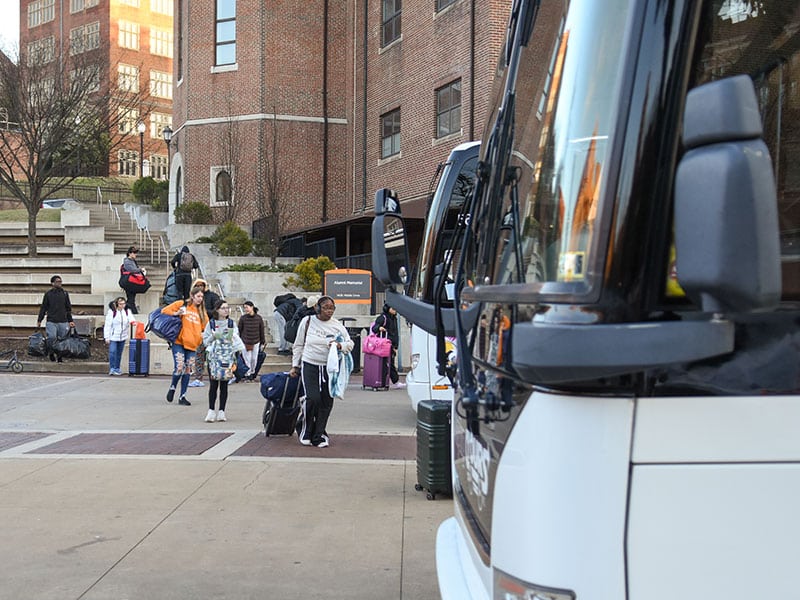 Students walk toward coach busses with luggage.