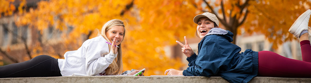 Two girls laying on their stomachs on the concrete. The girl on the left is waving her hand at the camera with a smile on her face. The girl on the right is holding up a peace sign and has an open mouth smile. This photo represents UT to West Tennessee.