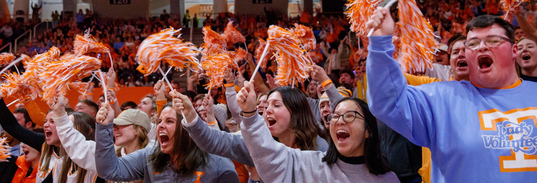 A group of diverse UT students join together to shake their orange and white pom poms at a UT Lady Vols game.
