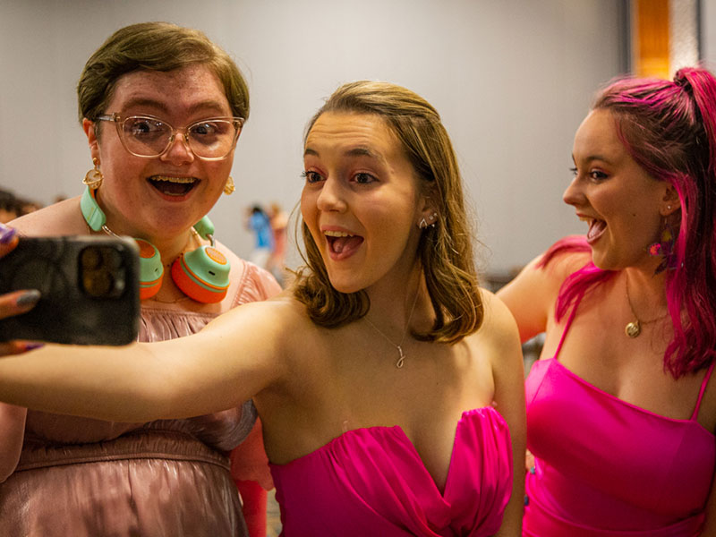 Three students wear pink formal dresses and pose for a selfie at Pride Prom.
