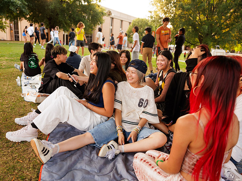 A large group of students playfully smoosh together on a picnic blanket in the HSS Plaza while many other students play games on the lawn behind them.