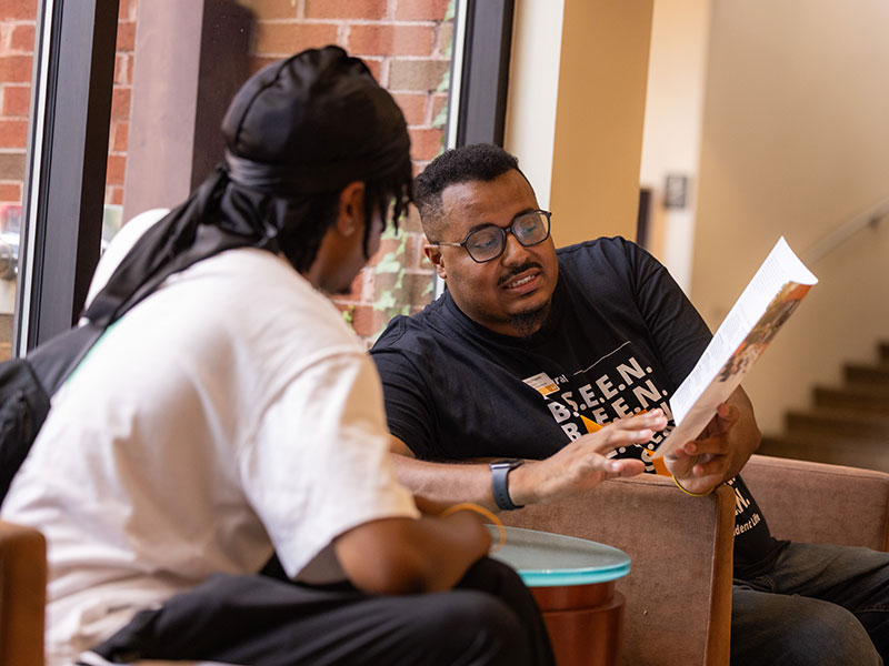A male student and staff member sit in the lobby of the Frieson Black Cultural Center conversing over a brochure.