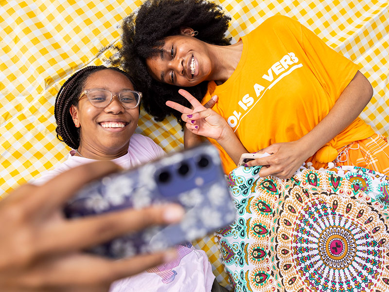 Two female students pose for a selfie against an orange and white checkered backdrop. One student wears an orange shirt with the Vol is a Verb name across the chest.
