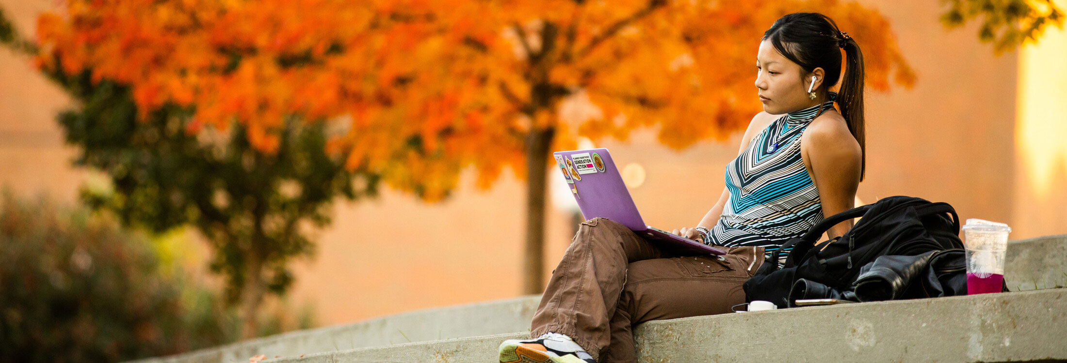 A student sits in the amphitheater on campus with orange fall leaves in the background and with their purple laptop on their lap. Their legs are crossed and they are wearing sneakers, brown pants and a striped blue blouse.