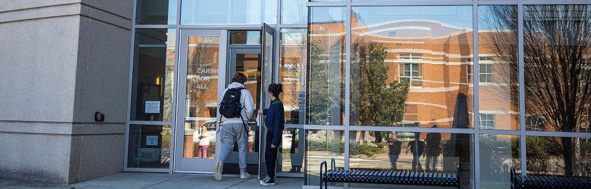An outdoor view of the student health center and one student is holding the door open for the other.