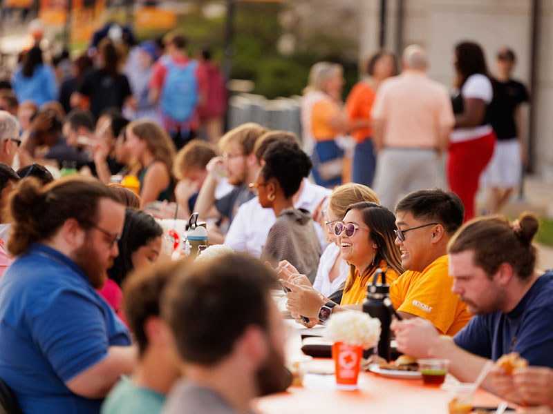 A long dining table on Pedestrian Walkway hosts large crowds of students gathered for a shared meal and conversation.