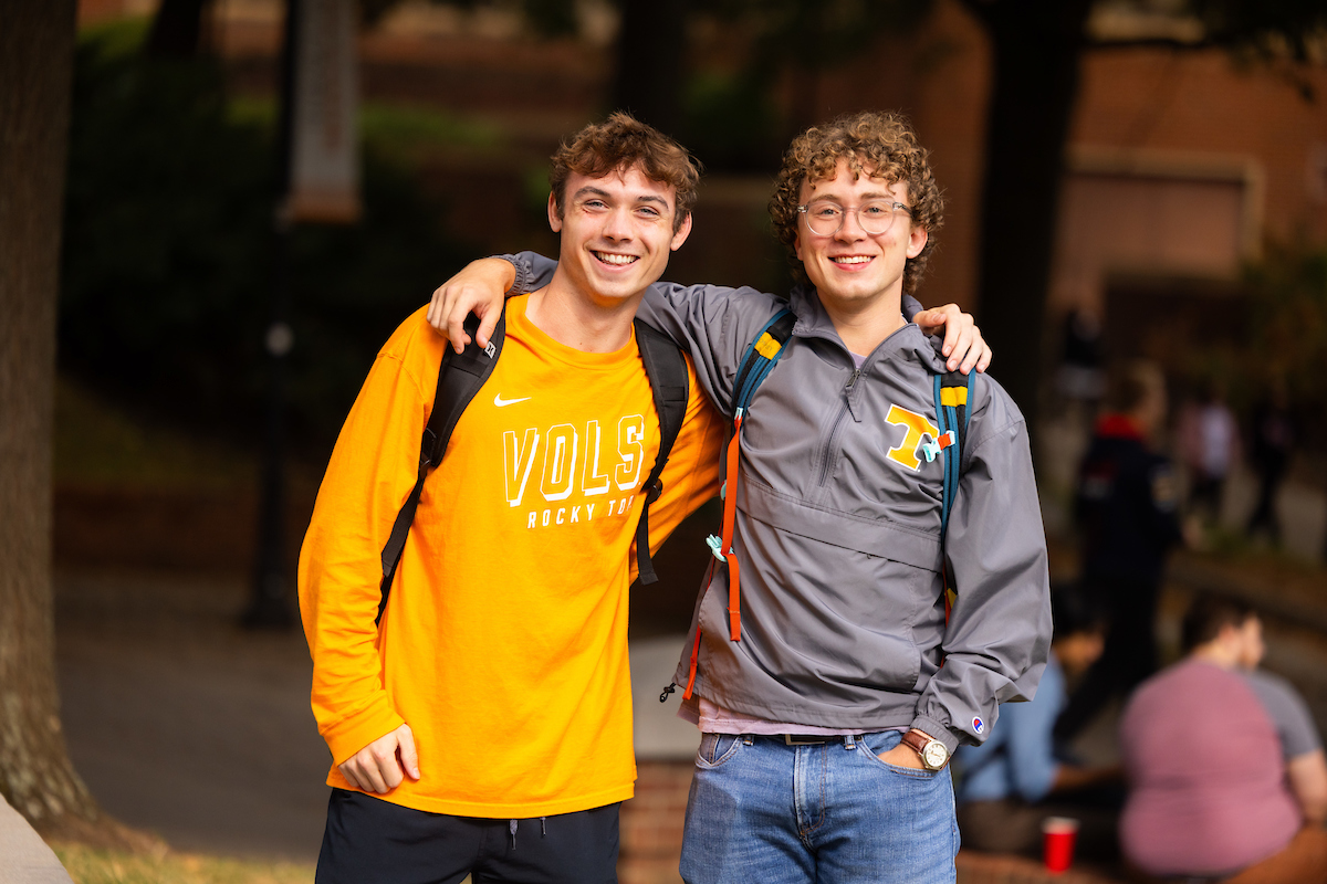 Two students stopped on PED Walkway for a photo. The student on the left is wearing an orange long sleeve t-shirt, while the one on the right is wearing a long sleeve gray Tennessee jacket.
