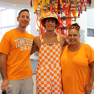 A family of three is smiling for a group photo inside the Student Union during Fall Family Weekend. They are all wearing orange clothing in support of the Tennessee Volunteers.
