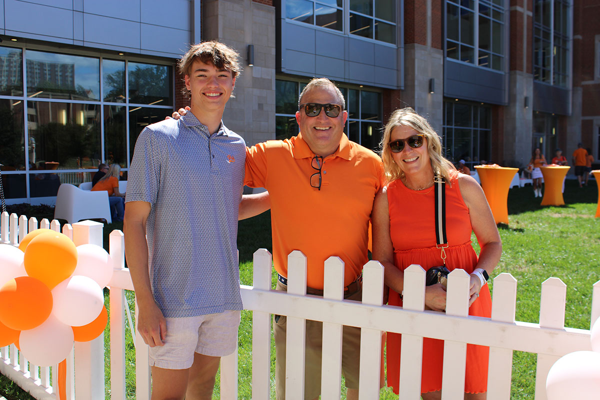 Three family members outside of the Student Union for Fall Family Weekend. Two members of the family are within the fence, while the other is outside of the fence. They have their arms around each other.