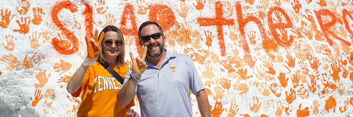 Two Volunteer parents standing in front of The Rock during the Slap The Rock event, which occurs during Fall Family Weekend. They are each holding up one of their hands, which has orange paint on it.