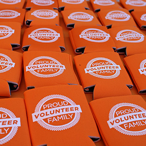 Rows of "Proud Volunteer Family" koozies laid out on a table for a family event. The koozies are orange with white lettering.