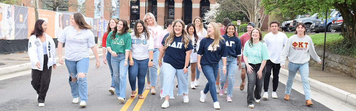Sorority and fraternity members at UTK are all walking together on the road in front of Neyland Stadium. They are smiling and laughing with each other.