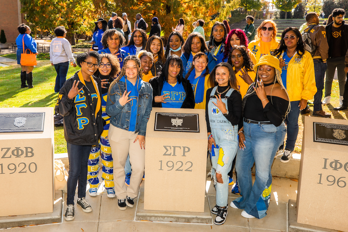Members of Sigma Gamma Rho Sorority, Inc. smiling for a group photo in front of the National Pan-Hellenic monuments. They are all wearing blue and yellow sorority apparel.