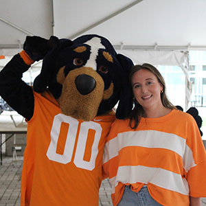 A staff member of the Office of Sorority & Fraternity Life smiling with the mascot, Smokey. Smokey is wearing an orange football jersey that has the number 00 on it.