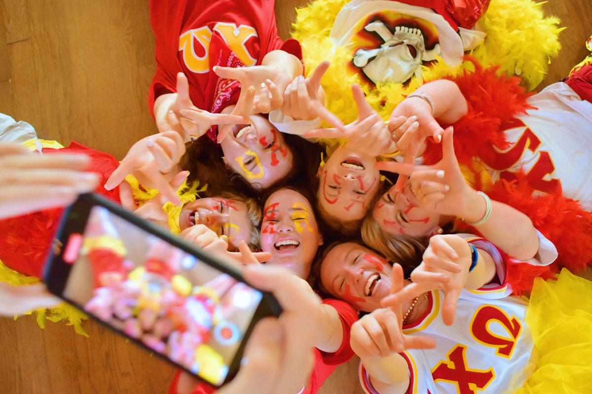 Several members of Chi Omega are laying on the floor with their heads all gathered together. They are all holding up a hand sign and smiling. They are wearing yellow, red, and white to represent their sorority colors. In the blurry foreground, someone is videoing them.