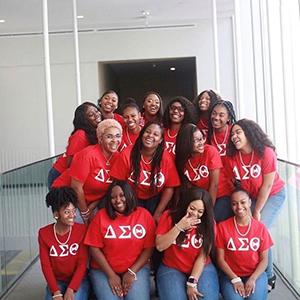 Members of Delta Sigma Theta Sorority, Inc. smiling for a group photo in the hallway. They are all wearing matching red shirts with white Greek letters on them.