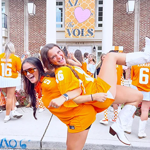Two local members of Delta Zeta posing outside of their sorority house within Sorority Village. One girl is on the other girl's back. They are both wearing orange Tennessee football jerseys.