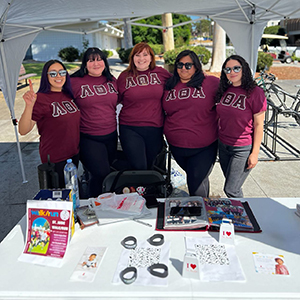Five members of Lambda Theta Alpha Sorority, Inc. during a philanthropy tabling event. They are all under a white tent.