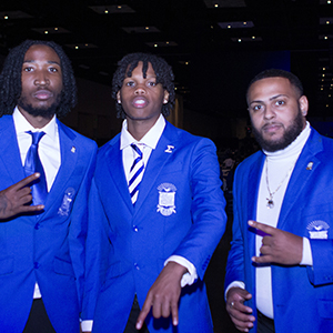 Three members of Phi Beta Sigma Fraternity, Inc. smiling with their hand sign. They are all wearing blue coats with the crest on the left hand side.