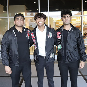 Three members of Sigma Beta Rho Fraternity, Inc. on a stage outside of the Student Union. They are all wearing a black bomber jacket with their Greek letters on it.