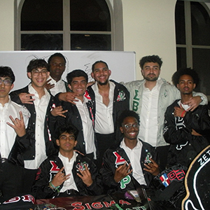 Various members of Sigma Beta Rho holding up their hand sign. They are inside of a building in front of a white board.