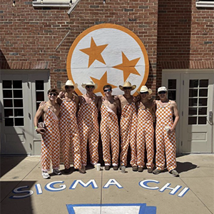 Seven local members of Sigma Chi in the back of their fraternity in front of the Tennessee Tri-Star wooden plaque. They are all wearing orange and white checkered overalls.