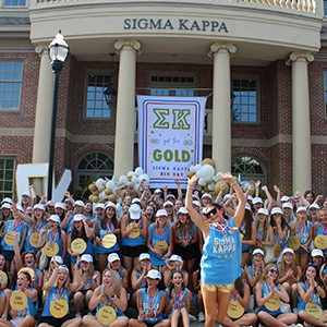 Several members of Sigma Kappa are standing outside of their sorority house on bid day. They are all wearing blue cutoff tank tops, and they have gold paper medals around their neck.