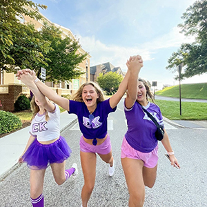Three local members of Sigma Kappa smiling in the street of Sorority Village. They are all holding hands and wearing different outfits that show their sorority pride.