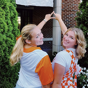 Two members of Delta Delta Delta in orange for Tennessee day during work week. They are outside of the sorority house. They are holding up a triangle with their fingers. Their heads are turned around their shoulder, but they are facing opposite of the camera.