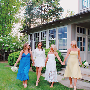 Four executive members of Delta Delta Delta outside of white and gray house. They are walking towards the camera and smiling at each other. 