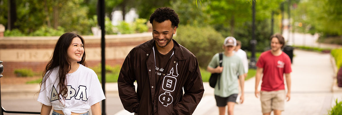 Two students that are apart of sorority and fraternity life are walking outside of fraternity park. The female student is wearing a white shirt with her sorority letters on it. The male student is wearing a long sleeve brown jacket with his fraternity letters on it.