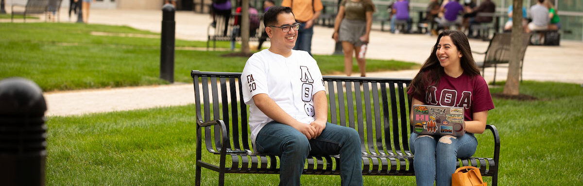 Two students within the Multicultural Greek Council are sitting on a bench outside of the Student Union. Both students are smiling. The male student is wearing a white shirt with his Greek letters on it. The female is wearing a maroon shirt with her Greek letters on it.