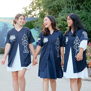 Three members of Delta Phi Lambda Sorority, Inc. walking and smiling at each other. They are all wearing baseball jerseys with their Greek letters on it.