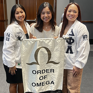 Three members of Delta Phi Lambda Sorority, Inc. at the Order of Omega event. Two members on the left and right are wearing white bomber jackets with their Greek letters on it. One member is holding up the Order of Omega banner.