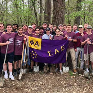 Dozens of members of Sigma Alpha Epsilon in the outdoors holding a purple and yellow flag with their Greek letters on it. Some men are holding shovels for their service event.
