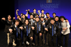 Members of the Lambda Phi Epsilon chapter on stage in the student union wearing Greek apparel and showcasing their hand sign. 