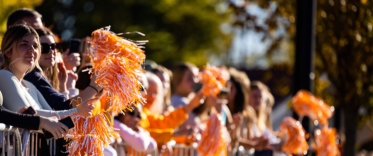 A photo of people standing behind a barricade, enjoying the parade with orange and white pom poms