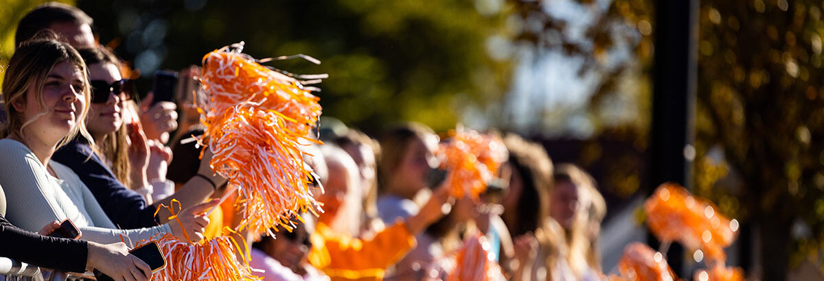 A photo of people standing behind a barricade, enjoying the parade with orange and white pom poms