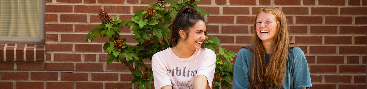 Two girls wearing t-shirts and smiling with the brick exterior of a residence hall behind them