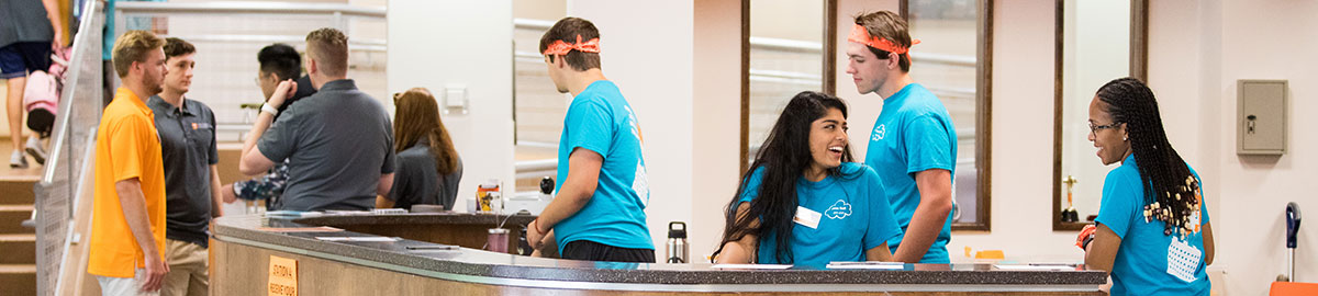 RAs standing behind the front desk in Hess Hall during move in, all wearing matching blue shirts