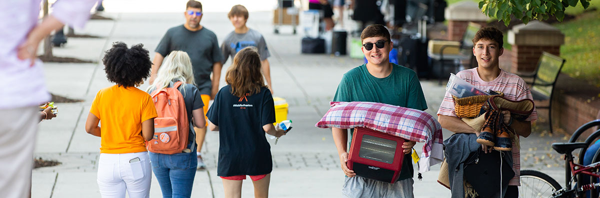 Two male students smiling as they have their arms full moving into Hess Hall on a sunny day