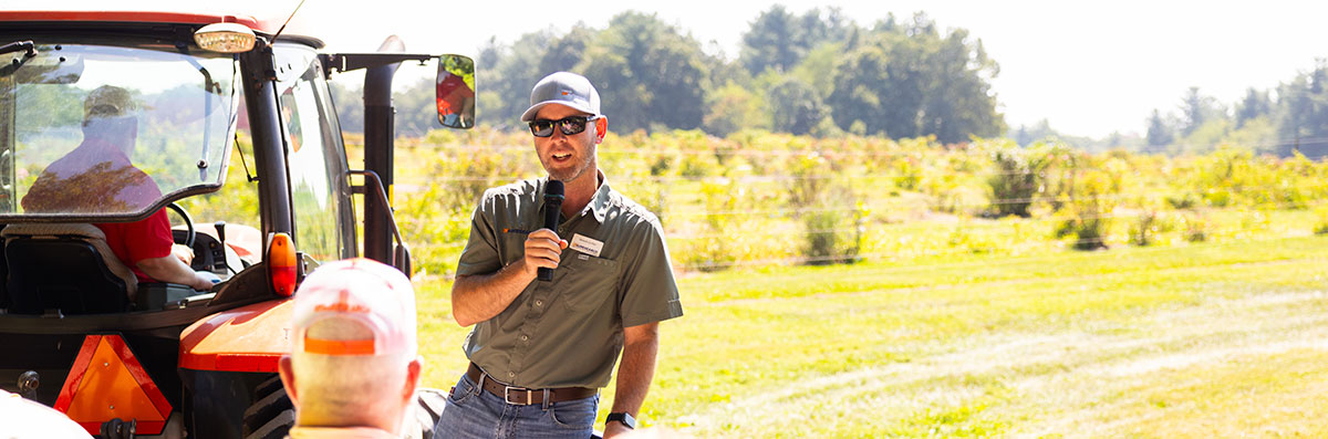 An agriculture student giving a tour to individuals on the back of a tractor. The student is wearing a button up and a gray UT hat