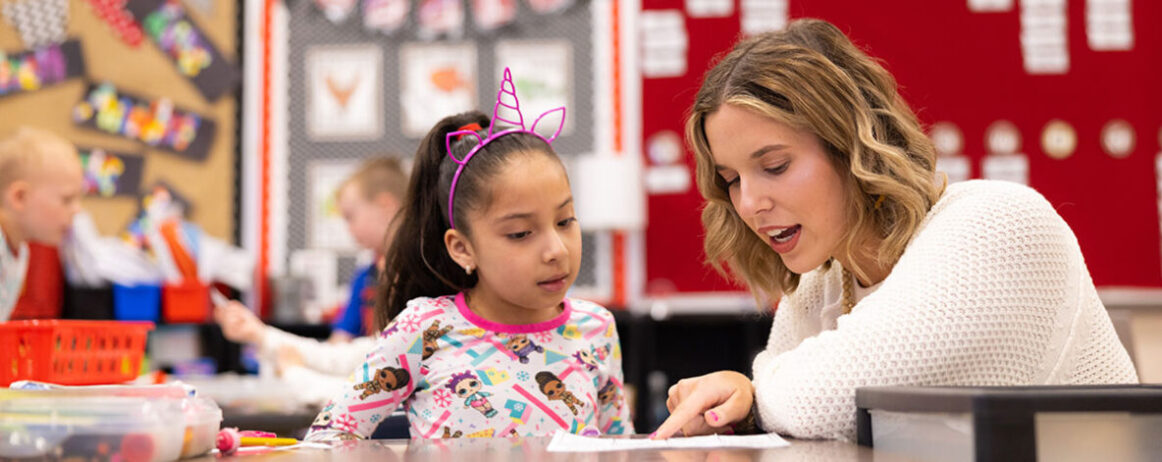 A student teaching an elementary school student who is wearing a pink unicorn headband