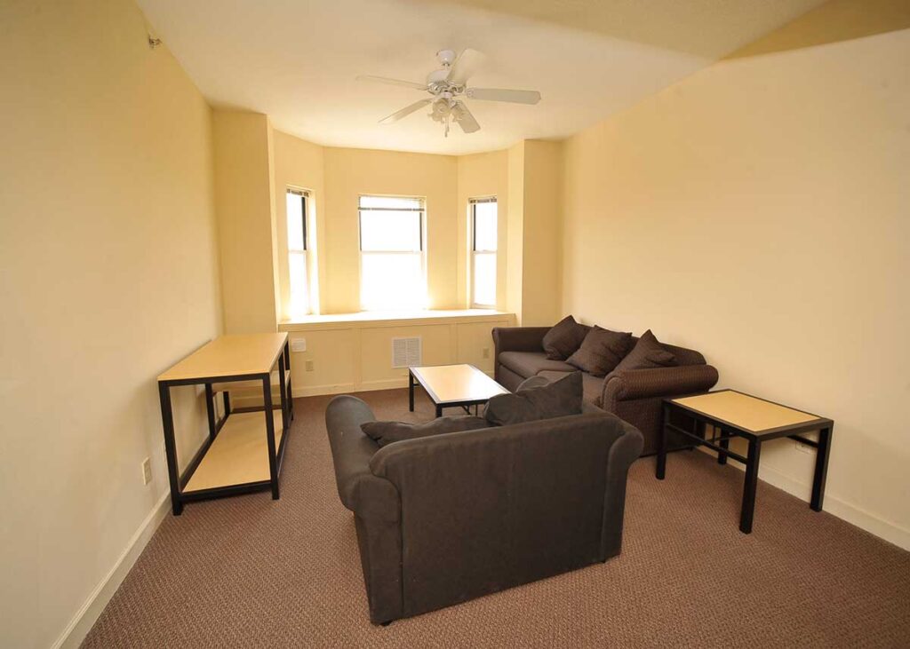 An undecorated living room with a cozy fabric armchair, three person couch, a side table, coffee table and entertainment stand with a bay window on the back wall