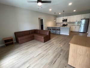 An undecorated living room with an L-shaped couch, one nightstand, three barstools a part of the kitchen island