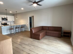 An undecorated living room with an L-shaped couch, one nightstand, three barstools a part of the kitchen island