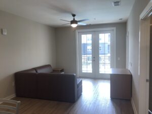 An undecorated living room with a television stand an L-shaped sofa with an end table and two french doors that lead to a balcony