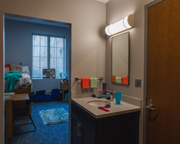 The bathroom sink in Magnolia Hall with the bedroom door opened showing the desk and bed