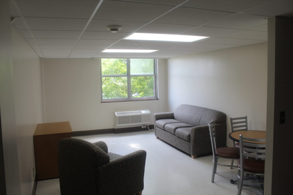 An undecorated living room with white tile floor, white ceiling tiles, a circular dining table with three visible chairs, one television console stand, three-seater couch and one chair.