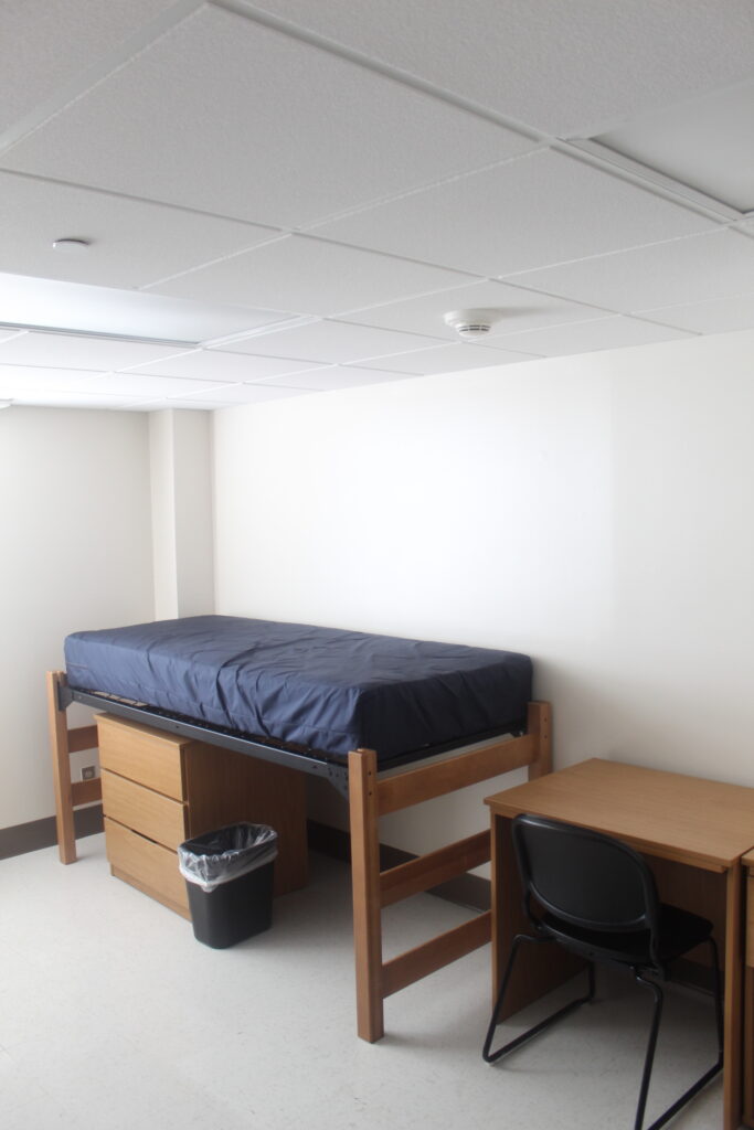 An undecorated bedroom with one twin XL bed that is slightly elevated with a three drawer dresser underneath it. There is also a small desk with a black chair to the right of the bed.